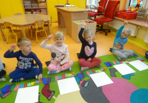 Czwórka dzieci siedzi na dywanie przed płytką i klockami Numicon, w prawej dłoni uniesiony mają pomarańczowy klocek.